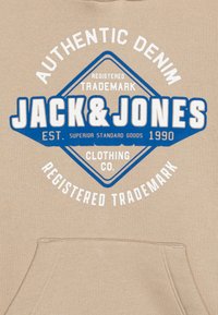 Bež dukserica s kapuljačom koja ima plavi dijamantni logo s bijelim tekstom. Prikazuje "JACK & JONES" i "AUTHENTIC DENIM" u podebljanom slovu. Mekana tekstura.