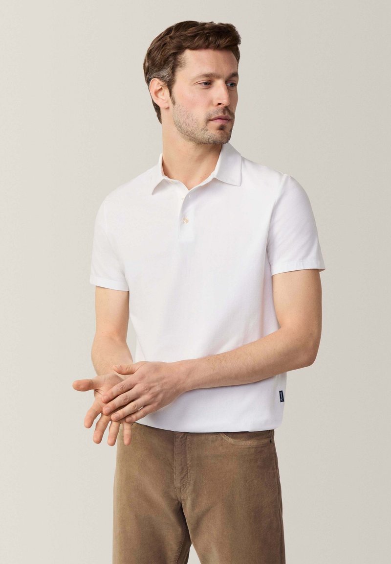Polo bianco a maniche corte in cotone, con colletto classico, abbottonatura a tre bottoni e logo discreto sull'orlo. Indossato con pantaloni marroni.