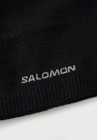 Čierna pletená čiapka s vyvýšeným logom "SALOMON" v bielej farbe. Tkanina vyzerá texturovane s hustou pletenou štruktúrou.