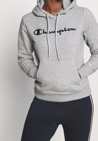 Person som bär en grå Champion-hoodie med fickor och marinblå byxor med röda och vita sidosträck, händerna i fickorna på hoodien.