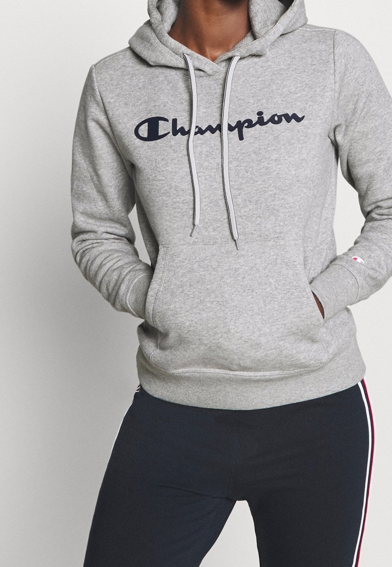 Person som bär en grå Champion-hoodie med fickor och marinblå byxor med röda och vita sidosträck, händerna i fickorna på hoodien.