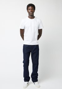 Witte katoenen T-shirt met korte mouwen, gecombineerd met donkerblauwe spijkerbroek en witte sneakers, staande tegen een effen achtergrond.