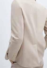 Blazer beige à coupe ajustée, présentant une texture lisse, une fente unique au dos et trois boutons décoratifs sur la manche.