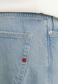 Lyseblå denimjeanslomme med synlige syninger og en lille rød rund nitte nær sømmen, båret med en hvid top.