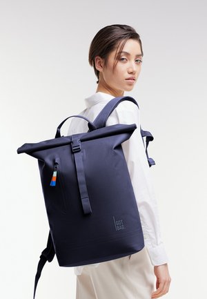 Persona joven con mochila enrollable azul marino con detalles de cremallera coloridos y logo "GOT BAG", vestido con camisa blanca y pantalones claros.