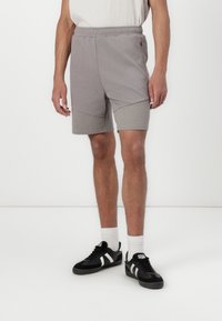 Un short de sport gris avec une taille élastique, présentant un panneau inférieur texturé et des poches latérales, porté avec des chaussures noires et des chaussettes blanches.