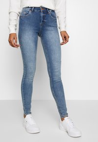 Skinny blue denim jeans with faded front, five pockets, and belt loops, paired with white sneakers and a light-colored sweatshirt.