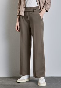 Wide-leg trousers in a muted brown fabric, featuring side pockets and a button closure. Paired with white sneakers for contrast.