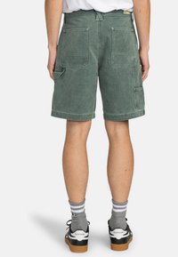 Shorts cargo verts en tissu doux et délavé ; dotés de deux grandes poches arrière et d'une coupe décontractée. Associés à des baskets noires et des chaussettes grises.