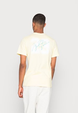 Mann trägt ein hellgelbes T-Shirt mit großer weißer "23" und einer blauen kursiven Unterschrift auf der Rückseite, dazu hellgraue Hose, blickt von hinten.