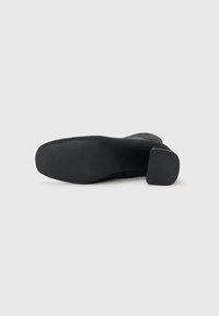 Black heeled shoe with a textured fabric upper, square heel, and rubber outsole featuring ridges for traction. Clear design and material details.