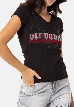 Kobieta ubrana w dopasowaną czarną koszulkę z dekoltem w serek, zdobioną czerwonymi i białymi cyrkoniami, w połączeniu z jasnoszarymi dżinsami.