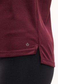 Κοντινό πλάνο στο τελειώμα πουκάμισου μπορντό με υφή και πλαϊνή σχισμή πάνω από μαύρο παντελόνι, με μικρό ανακλαστικό λογότυπο κοντά στην κάτω άκρη.