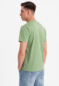Camiseta verde de manga corta, con un patrón moteado. Textura suave, escote redondo, exhibida con jeans desgastados de color azul claro.