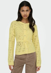 Cardigan jaune en maille avec un motif en dentelle complexe, boutons en forme de cœur et taille à cordon. Manches longues, tissu texturé.