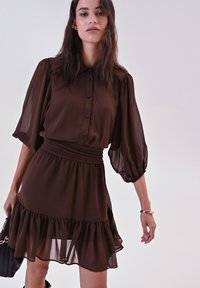 Donna che indossa un vestito trasparente marrone scuro con maniche a palloncino e orlo arricciato, mentre tiene in mano una borsa nera, in piedi contro uno sfondo chiaro e semplice.