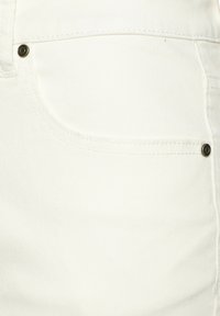 Tela de mezclilla blanca con un bolsillo frontal que cuenta con un remache de metal, textura suave y un sutil patrón de sarga. Detalles de costura a lo largo de las costuras.