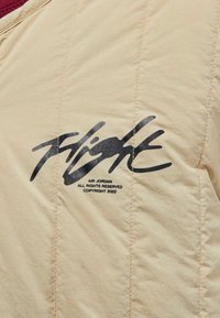 Beige quiltet jakke med en bordeaux krave. Sort "Flight" logo med brandingdetaljer trykt på brystet. Struktureret stoffinish.