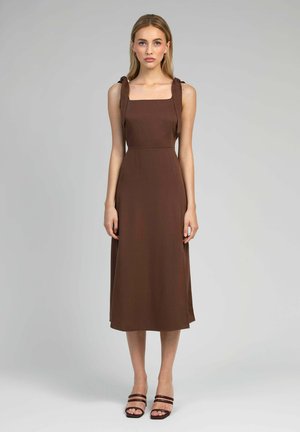 Femme debout en robe midi marron sans manches avec un décolleté carré et des bretelles à nouer aux épaules, portant des sandales à talons ouvertes.