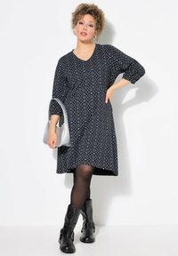 Schwarzes gemustertes Kleid mit drei Viertelärmeln, V-Ausschnitt und leicht ausgestellt geschnittenem Saum. Trägt schwarze Stiefel und eine silberne Handtasche.