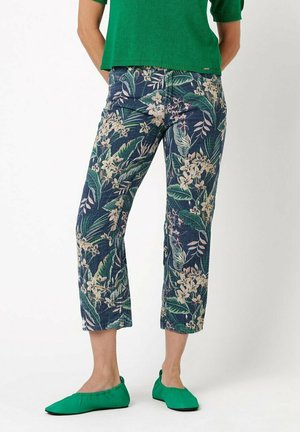 Person trägt gekürzte Hose mit Blumenmuster, grünes Oberteil und passende grüne flache Schuhe und steht vor einem schlichten weißen Hintergrund.