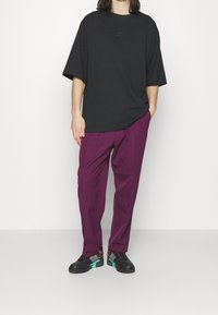 Černé oversized tričko s krátkým rukávem a logem Nike, v kombinaci s volnými fialovými kalhotami a černými teniskami s tyrkysovými a růžovými detaily.