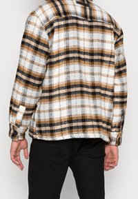 Man som bär en brun, svart och vit rutig kavaj med svarta byxor, sedd bakifrån mot en enkel ljus bakgrund.