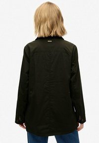 Chaqueta ligera de color verde oscuro con cuello, mangas largas y puños abotonados. Presenta un detalle de etiqueta en la parte posterior y un diseño de corte recto.