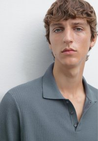 Szara prążkowana koszulka polo z kołnierzykiem, z trzema guzikami i teksturowaną tkaniną. Model ma neutralny wyraz twarzy.