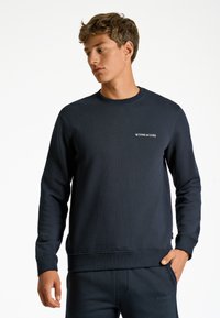Mörkblå sweatshirt i en strukturerad tygkvalitet, med en rundad halsringning, ribbade ärmslut och en liten vit logga på bröstet.