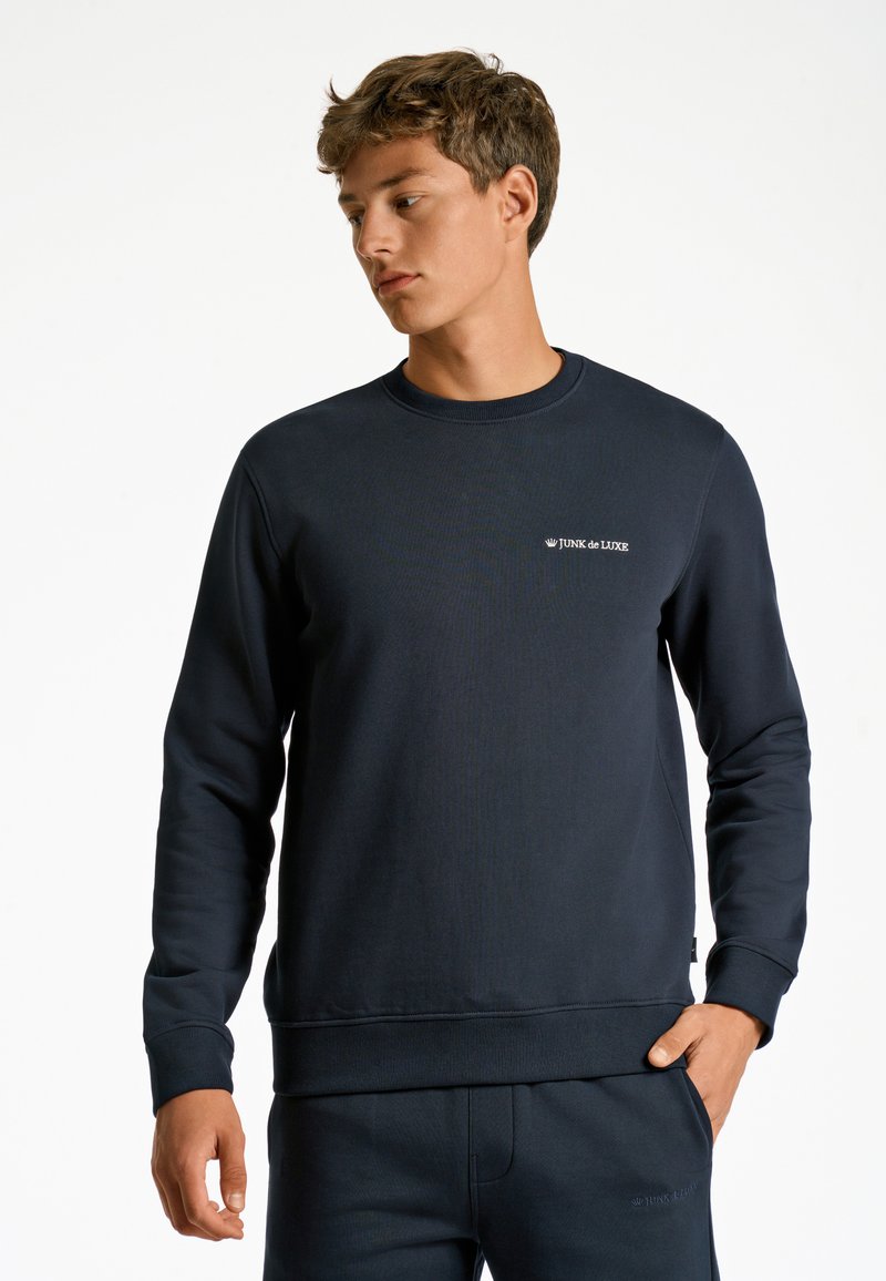 Mörkblå sweatshirt i en strukturerad tygkvalitet, med en rundad halsringning, ribbade ärmslut och en liten vit logga på bröstet.