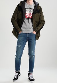 Oliwkowo-zielona kurtka z czarnymi akcentami, szara bluza z nadrukiem graficznym, postrzępione niebieskie dżinsy oraz czarne sneakersy za kostkę.