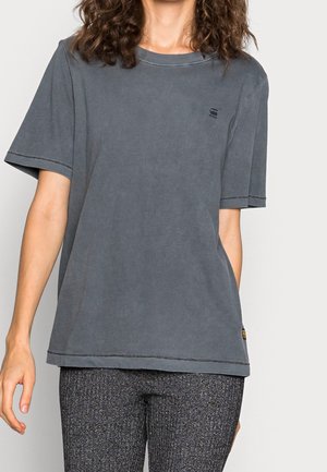 Kvinde iført en løs, falmet grå t-shirt med korte ærmer og teksturerede mørkegrå bukser, stående foran en ensfarvet baggrund.