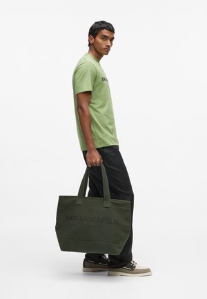 Man in green shirt and black pants carrying large olive tote bag with "KARL LAGERFELD" print, standing against white background.
