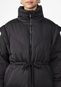 Svart polstret jakke med høy krage, glidelås foran, elastisk midje med snor, og kviltet søm. Glatt tekstur, minimalistisk design.