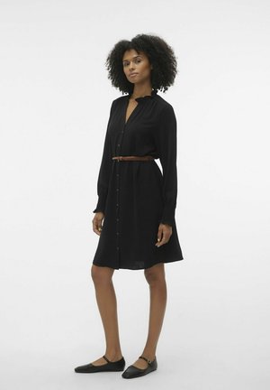 Robe noire à manches longues et avec un col boutonné, resserrée à la taille par une ceinture en cuir tressé marron. Portée avec des chaussures plates noires.