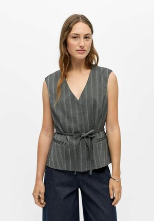 Femme aux cheveux châtain clair portant un haut gris sans manches à fines rayures avec une ceinture nouée à la taille et un pantalon large foncé, sur fond blanc.