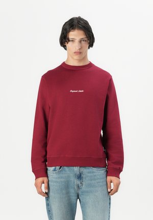 Burgunderfarget sweatshirt laget av bomull, med rund hals og ribbede mansjetter. Framsiden har hvit brodering med teksten "Original Standard."