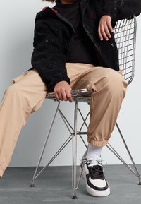 Czarna polarowa kurtka, czarna bluzka z długim rękawem, beżowe spodnie z tapered, oraz czarno-białe sneakersy z akcentami w postaci sznurówek. Siedzi na metalowym krześle.