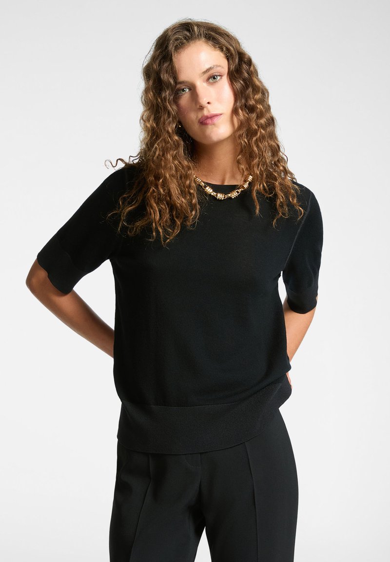 Pull noir à manches courtes avec une coupe décontractée et un ourlet côtelé. Texture douce avec un col rond, associé à un collier métallique.