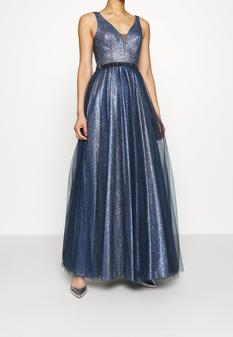 Femme portant une robe longue sans manches bleu marine scintillante avec un profond décolleté en V et une ceinture noire à perles à la taille, associée à des talons pointus argentés.