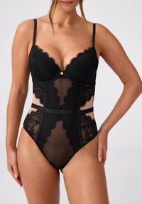 Czarne koronkowe body z przezroczystymi panelami i falistymi krawędziami, z regulowanymi ramiączkami i małym metalowym akcentem na środku przodu.