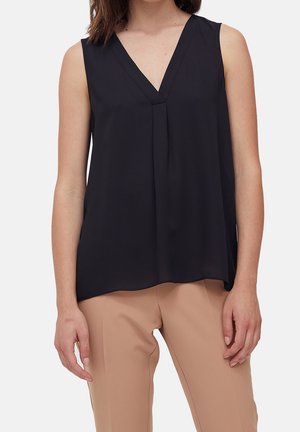 Hihaton musta V-kaula-aukkoinen paita, jossa edessä laskos, yllään beige räätälöidyt housut seisovalla henkilöllä.