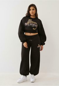 Fekete cropped pulóver leopárdmintás hímzéssel és "True Religion" logóval, fekete melegítőnadrággal és fehér sportcipővel párosítva.