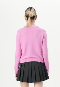 Vrouw met blond haar in een knot, gekleed in een roze lange mouwen trui en een zwarte geplooide rok, staande tegen een witte achtergrond.