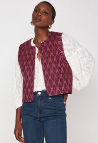 Donna che indossa orecchini a cerchio d'oro, una blusa bianca testurizzata con maniche a sbuffo, un gilet senza maniche a fantasia viola e jeans blu scuro.