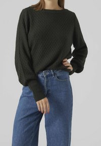 Maglione lavorato a maglia verde scuro con un motivo testurizzato, maniche larghe e polsini a coste, abbinato a jeans in denim blu a vita alta.