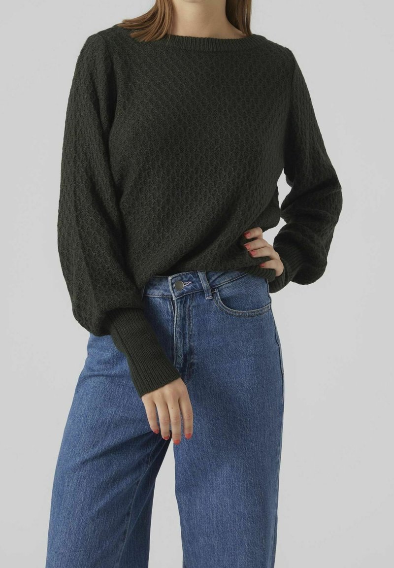 Maglione lavorato a maglia verde scuro con un motivo testurizzato, maniche larghe e polsini a coste, abbinato a jeans in denim blu a vita alta.