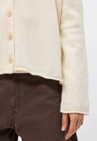 Κρεμιστός πλεχτός κάρντιγκαν με στρογγυλή λαιμ�όκοψη, πέντε κουμπιά και ριμπέ μανίκια, σε συνδυασμό με σκούρο καφέ παντελόνι. Ορατή υφή και ραφές.