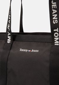Sac fourre-tout noir en tissu durable, avec des poignées en textile doubles arborant le logo "Tommy Jeans" et un détail de couleur emblématique au centre.